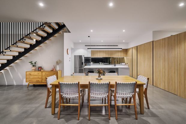 A bright, open-plan contemporary kitchen and dining area with a large timber dining table, woven dining chairs, grey concrete-look flooring, extensive vertical timber cabinetry, and a sleek island kitchen. A striking open-riser staircase with black metal structure and timber treads runs along the left side, while recessed ceiling lights provide even illumination.