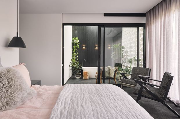 A contemporary bedroom with a soft, neutral palette, featuring a low-profile bed dressed in a white quilt and textured pillows, a black pendant light, dark woven lounge chairs, and full-height sheer curtains. Black-framed glazed sliding doors open to a planted enclosed courtyard or sunroom with ribbed dark wall cladding and a decorative breeze-block screen.