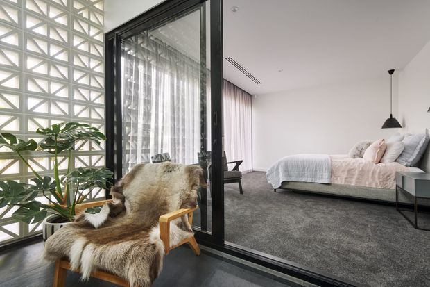 A contemporary bedroom viewed through a black-framed glazed partition, with a low platform bed, soft neutral bedding, grey carpet, sheer curtains, and a black pendant light. In the foreground, a timber-framed armchair is draped with a sheepskin throw beside a leafy indoor plant. A patterned concrete breeze-block screen provides a distinctive architectural feature along the left side.
