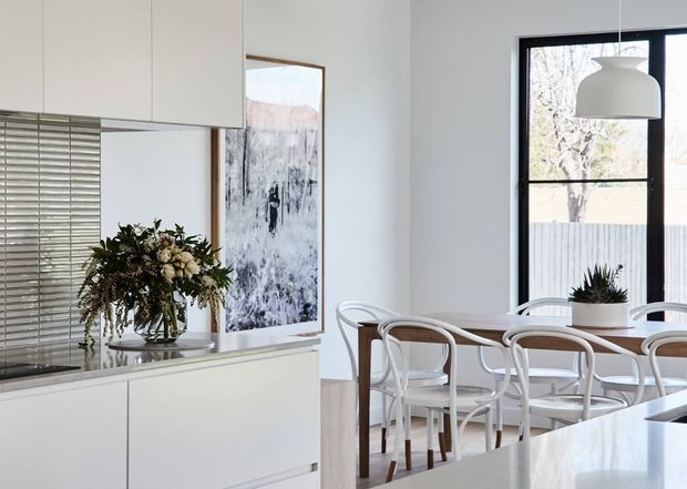 A bright, refined open-plan dining and kitchen interior. White flat-panel cabinetry and a pale benchtop occupy the foreground, with a timber dining table surrounded by white bentwood-style chairs beside large black-framed windows. A white pendant light, venetian blinds, artwork, and a floral arrangement complete the space.