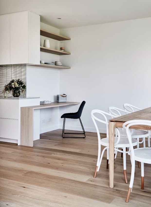 Bright open-plan kitchen and dining area with white flat-panel cabinetry, a subtly textured tiled splashback, floating timber shelves, a compact timber study nook with a black chair, and a timber dining table surrounded by white chairs. The room has light timber flooring and a clean, minimal aesthetic.