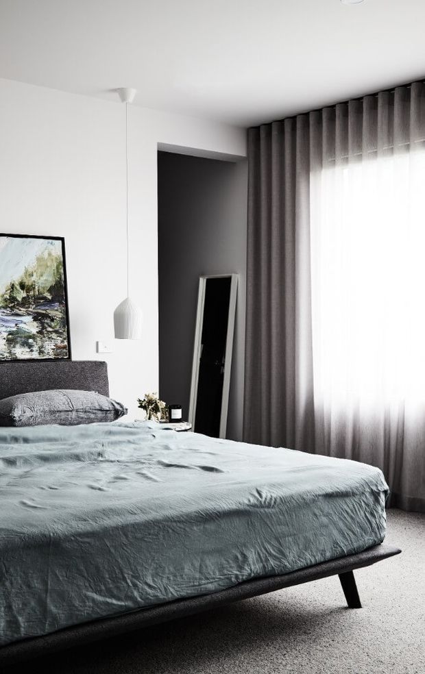 A contemporary bedroom with a low-profile upholstered bed dressed in pale blue-grey bedding, a dark textured headboard, white walls, charcoal floor-to-ceiling curtains, and a large bright window. A slim pendant light, leaning full-length mirror, artwork, and small bedside decor complete the restrained monochromatic scheme.