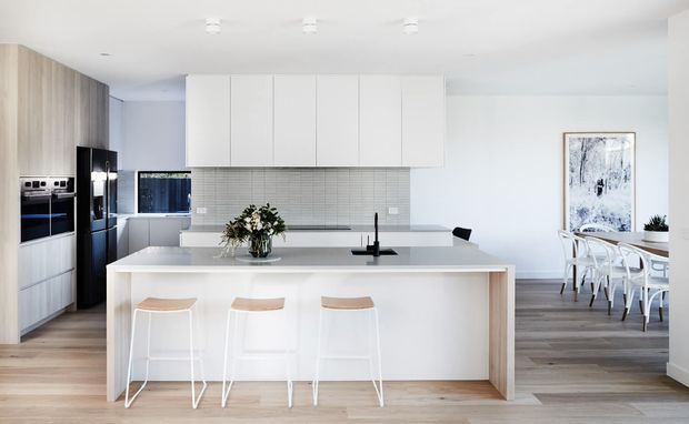 Bright open-plan kitchen with a large white island and three stools, pale timber flooring, white flat-panel cabinetry, grey tiled splashback, integrated dark appliances, and a dining area visible beyond. The space has a clean, contemporary minimalist feel with abundant natural light.