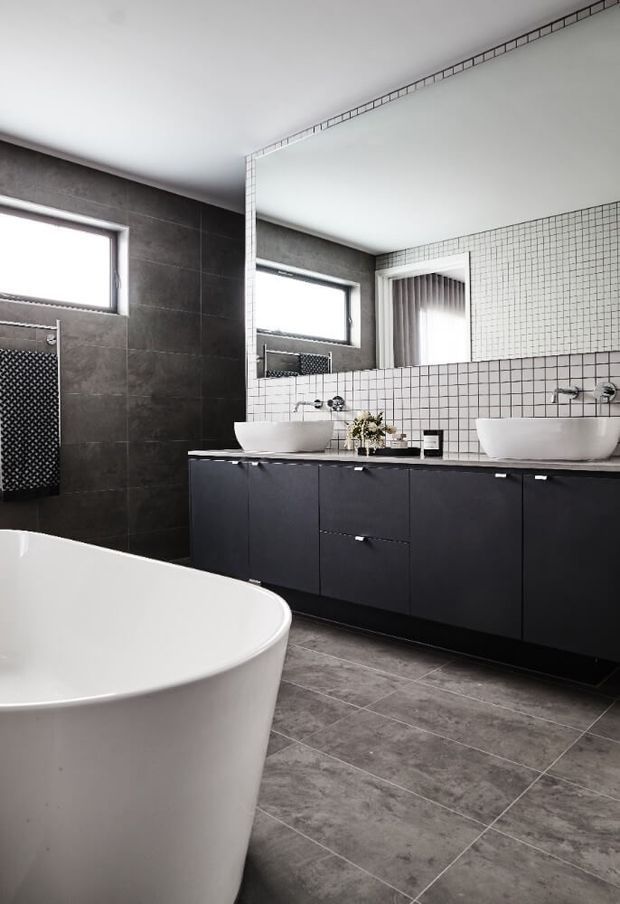 Contemporary bathroom featuring a freestanding white bathtub, double white vessel basins on a long matte-black vanity, dark large-format wall tiles, grey floor tiles, and a white small-grid tiled backsplash and mirror surround. A high horizontal window and shower area are visible in the reflection.