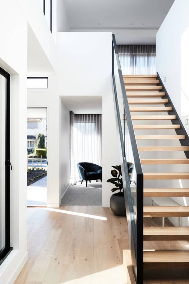 Bright, minimalist residential stairway with an open-riser timber staircase, slim dark metal and glass balustrade, pale timber flooring, white walls, and full-height sheer curtains. A small seating nook and potted plant are visible beside the stair, with strong natural light entering through large windows and an open doorway.