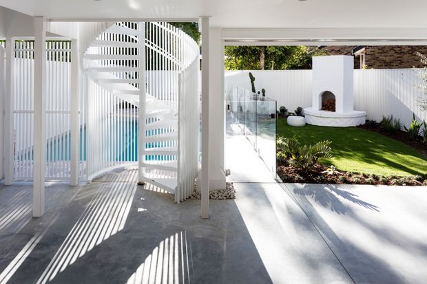 Bright contemporary outdoor entertaining area featuring a white spiral staircase enclosed by vertical white slatted screening, polished concrete flooring, and a view into a landscaped courtyard. The courtyard includes a glass pool or balustrade enclosure, manicured lawn, white rendered feature fireplace, and patterned masonry boundary wall.