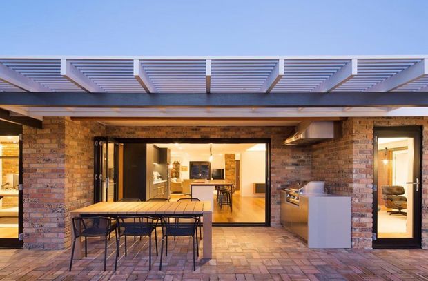 A contemporary brick-clad outdoor entertaining area with a covered pergola and exposed steel structure. The paved patio contains a dining table and chairs, while a built-in outdoor kitchen sits to the right. Large glazed openings connect the terrace to a bright interior kitchen and living spaces.