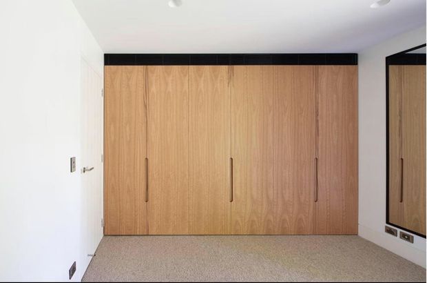 Minimal, modern built-in wardrobe spanning the far wall of a compact room. The wardrobe has tall light timber-grain doors with slim vertical handles, a black horizontal header and matching black-framed full-height mirror on the right wall. White walls and ceiling, recessed lighting, electrical outlets, and a neutral carpeted floor create a clean, understated interior.