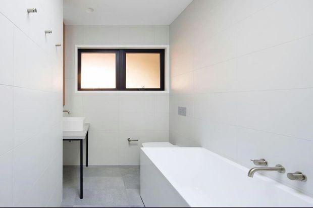 Bright, minimalist bathroom with a white built-in bathtub, pale tiled walls and floor, slim dark-framed horizontal window, and a compact vanity with black metal legs. The space has clean contemporary lines and restrained fixtures.