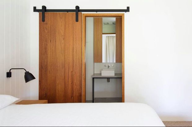 A minimalist bedroom with a white bed and black wall-mounted reading lamp, featuring a warm timber sliding barn door opening directly to a compact bathroom. The bathroom includes a wall-mounted vanity with a white basin, dark countertop, mirror, and curtain-covered window. Neutral white walls and understated industrial-style hardware create a clean, calm aesthetic.