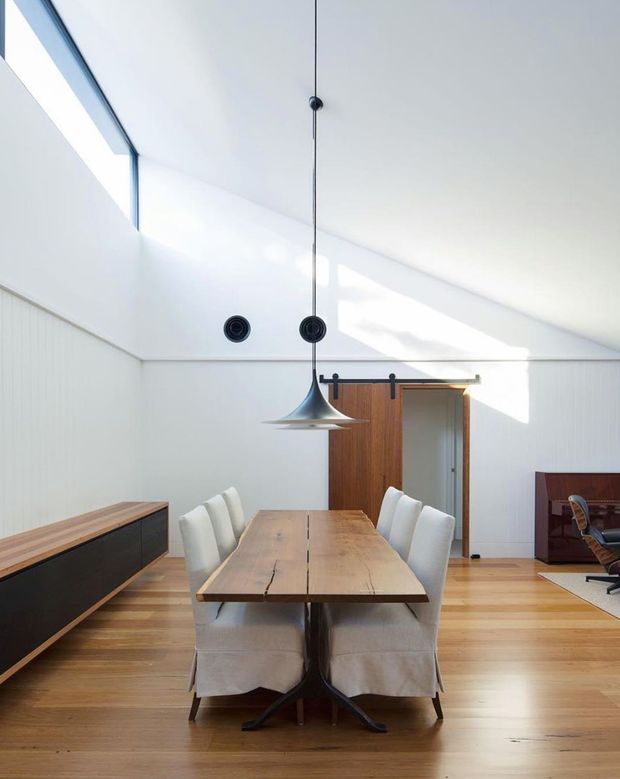 Bright, minimalist dining room with a long natural timber dining table surrounded by upholstered white chairs. The room features white vertical wall panelling, warm timber flooring, a sculptural suspended pendant light, a timber sliding barn-style door, and high angled ceilings with clerestory-like glazing. A low dark timber sideboard and part of an adjacent study or living area are visible.