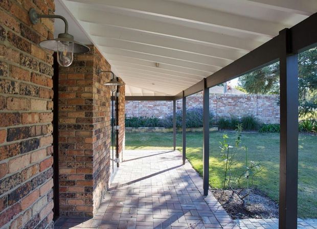 Covered brick-paved verandah running alongside a brick house, with exposed brick walls, dark slender support posts, a white-lined pitched roof soffit, and wall-mounted industrial-style lights. The verandah overlooks a landscaped lawn and garden enclosed by a brick boundary wall.
