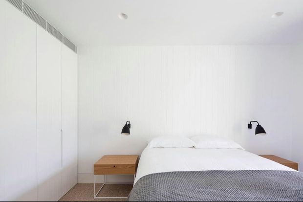 A bright, minimalist bedroom with a white upholstered or panelled wall, integrated floor-to-ceiling white cabinetry, a low bed dressed in white bedding, pale wood bedside tables, black wall-mounted reading lamps, and a textured grey throw. The room has a clean, restrained contemporary appearance.