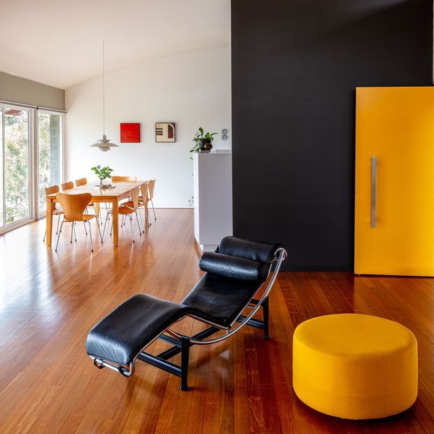 A bright, open-plan mid-century-inspired interior with polished timber flooring, a black leather lounge chair and ottoman in the foreground, and a compact dining area beside full-height glazing. A bold yellow pivot-style door and matching artwork provide strong colour accents against a dark feature wall and white walls.