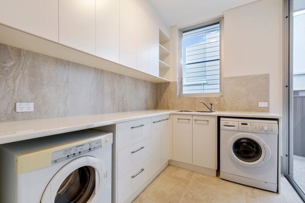 Bright, compact modern laundry with extensive white built-in cabinetry, pale stone-look benchtops and splashback, two front-loading washing appliances, a sink, tiled flooring, and a window providing natural light.