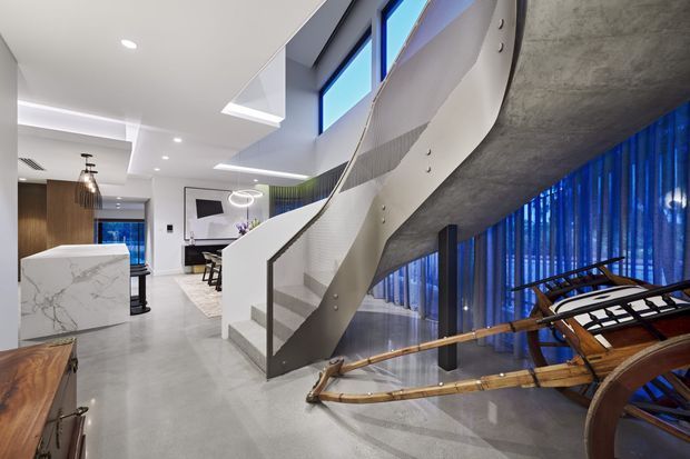 A contemporary open-plan interior centered on a sculptural curved staircase with white perforated balustrades and grey concrete-like surfaces. The space includes a polished grey floor, white ceiling with recessed lighting, dark timber cabinetry, a kitchen island with a marble-look benchtop, blue sheer curtains, and a distinctive wooden rowing boat displayed beneath the stair.
