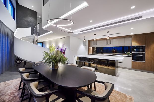 A high-end open-plan dining and kitchen interior with a large dark oval dining table and upholstered chairs in the foreground. The space features a white sculptural spiral staircase, concealed integrated kitchen cabinetry in timber and white finishes, a long island with bar seating, dark blue reflective splashback, pendant lighting, recessed ceiling lights, and pale polished flooring.