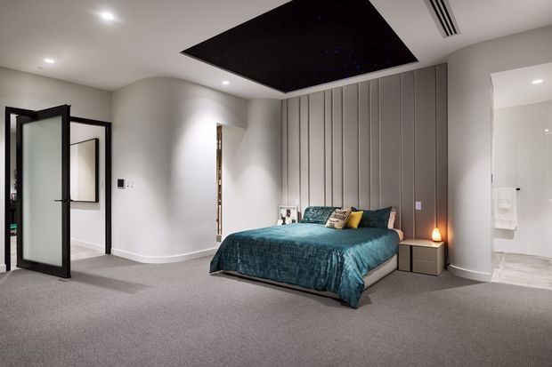A spacious contemporary bedroom with a low upholstered bed dressed in deep teal bedding and accent cushions, flanked by simple bedside tables. The room has grey carpet, pale curved walls, full-height vertical-panel cabinetry behind the bed, recessed lighting, a dark star-like ceiling feature, and an open black-framed glass door leading toward an ensuite bathroom.