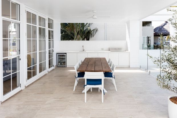 Bright, open-plan covered outdoor entertaining area with a long timber dining table and white chairs, a compact white kitchenette with sink and under-counter beverage fridge, extensive white-framed glazed doors, pale tiled flooring, ceiling fan, and tropical foliage visible beyond. The space has a clean coastal feel and connects visually to an exterior courtyard or garden.