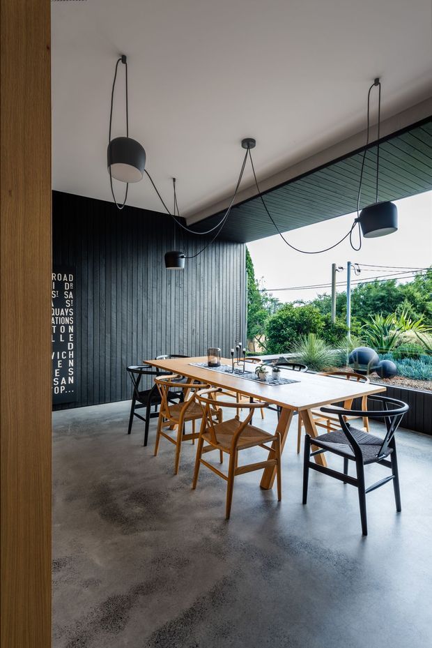 A contemporary dining area beneath a covered outdoor or semi-enclosed terrace. A long light timber dining table is surrounded by mixed timber and black-framed chairs. The space features a polished concrete floor, vertically grooved dark wall cladding, a dark slatted ceiling, and multiple black pendant lights with exposed cords. Large open glazing or an opening looks onto landscaped greenery and neighboring utility lines. A narrow black typographic wall artwork is partially visible at the left.