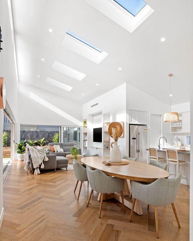 Bright open-plan interior combining a living room, dining area, and kitchen. The room features white walls and ceiling, multiple skylights and recessed downlights, extensive glazing, warm timber herringbone flooring, a large rounded-edge timber dining table with upholstered chairs, a grey sectional sofa, and white kitchen cabinetry with integrated appliances. Indoor plants and natural light create an airy contemporary atmosphere.