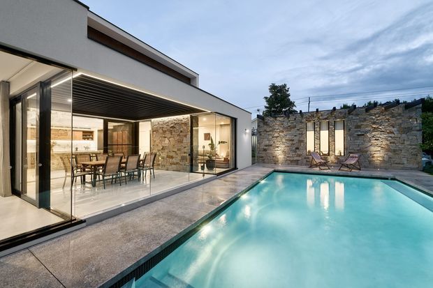 Contemporary residential courtyard viewed from outdoors, featuring a crisp white rendered pavilion with extensive floor-to-ceiling glazing, a covered outdoor dining area, and a large rectangular swimming pool. A textured natural-stone boundary wall with narrow vertical windows forms a strong backdrop, while integrated linear lighting adds a refined evening atmosphere.