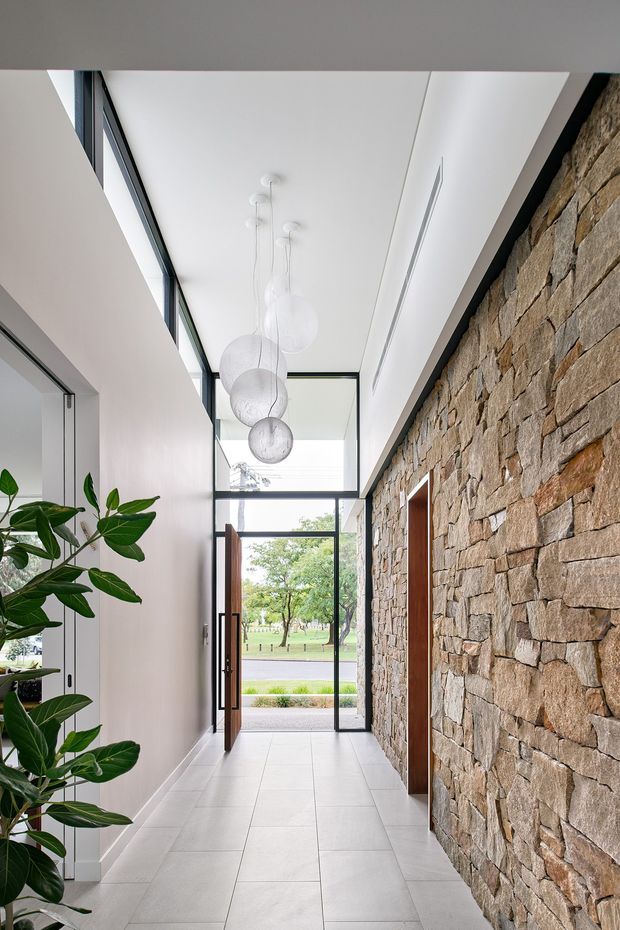 A bright, narrow contemporary entry hall with a long exposed natural stone feature wall on the right, smooth white walls and large pale floor tiles. Full-height black-framed glazing and a timber-framed open front door bring in views of greenery, while a cluster of white pendant lights hangs from the high ceiling. A leafy indoor plant occupies the left side.