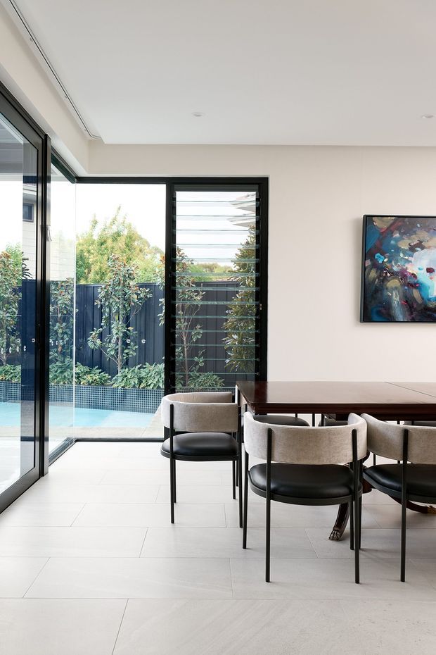 Bright contemporary dining area with a dark timber dining table and upholstered black-and-beige chairs, opening through full-height black-framed glazing to a small tiled courtyard and pool. A colourful abstract artwork hangs on the light neutral wall, and a horizontal louvred window provides ventilation and garden views.