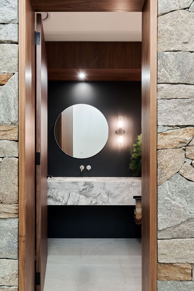 A compact, sophisticated bathroom viewed through a timber-framed doorway. The room features a black wall with a large round mirror, warm wall lighting, a floating marble vanity with wall-mounted taps, pale tiled flooring, and dark timber joinery. The surrounding doorway and exterior wall are clad in rough stacked natural stone.
