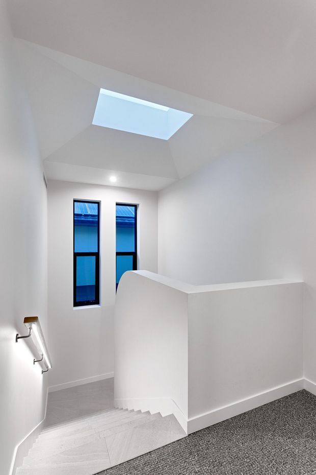 Bright minimalist stairway descending to a lower level, with white walls, a white balustrade, pale tiled steps, grey carpet at the landing, narrow blue-tinted windows, and a rectangular skylight illuminating the stairwell.