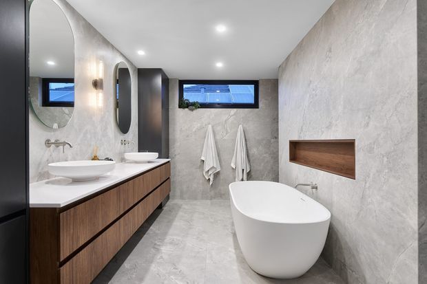 Contemporary bathroom with a freestanding white bathtub, double vessel basins on a dark timber vanity, large rounded mirrors, pale stone-look wall and floor finishes, recessed wall niche, and a narrow high window. Recessed ceiling lighting creates a clean, refined atmosphere.