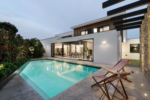 Exterior architectural view of a contemporary multi-level home with crisp white rendered walls, extensive dark-framed glazing, projecting horizontal roof or sunshade elements, and a textured stone-clad wall. A turquoise swimming pool and paved pool terrace occupy the foreground, with an open-plan dining area visible through the glazing.