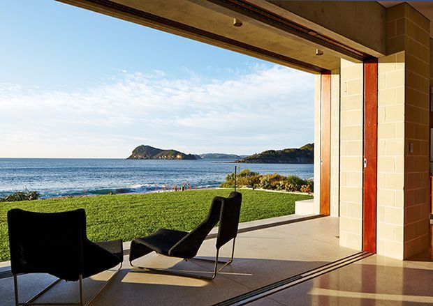A modern open-plan interior opening directly onto a waterfront lawn and ocean view. The frame is dominated by a wide glazed sliding-door opening with exposed track, pale tiled masonry walls, a timber-toned ceiling and trim, and polished neutral flooring. Two dark lounge chairs sit in the foreground, while a coastal bay and headland are visible beyond the lawn.