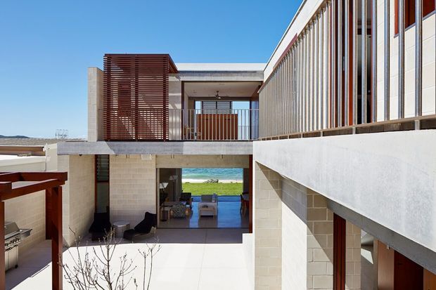 A contemporary multi-level coastal residence organized around an open courtyard. Pale masonry walls and concrete slabs frame a sheltered outdoor living area, while timber screening, vertical fins, and expansive glazing create layered privacy and strong horizontal and vertical lines. Through the opening, a bright blue ocean and sandy shoreline are visible beyond the house. An outdoor kitchen or barbecue area appears at lower left.
