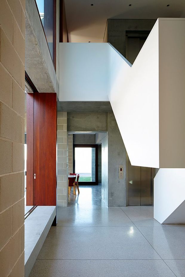 A tall, modern interior hallway or entry volume with a polished light-grey concrete or terrazzo floor, exposed concrete walls and beams, pale blockwork, and bold white angular stair or balustrade forms. A large vertical timber door or panel adds warmth on the left, while a dining area is glimpsed at the far end. The space is bright and architectural, with strong geometric lines and a multi-level circulation layout.