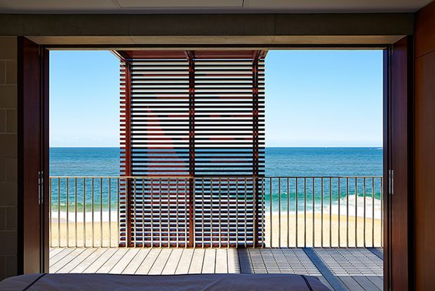 View from a bedroom through wide open timber-framed glazing onto a balcony or deck overlooking a sandy beach and ocean. A prominent external louvred screen provides sun shading and casts a striped shadow across the pale timber decking; dark interior wall and ceiling framing surround the opening.