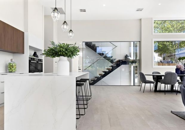 Bright, open-plan contemporary kitchen and dining area with a large white stone island, dark wood overhead cabinetry, integrated appliances, pendant lighting, and black bar stools. Glazed partitions reveal a modern staircase with dark treads, while a dining table and upholstered chairs sit beside large windows overlooking greenery.