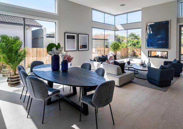 Bright, open-plan contemporary living and dining area with extensive floor-to-ceiling glazing and a high ceiling. A dark oval dining table with upholstered chairs occupies the foreground, while a light sectional sofa, dark lounge chairs, coffee tables, and large artwork define the living area. The room opens visually to a landscaped outdoor space and is finished in pale neutral tones with dark furnishings and blue accents.