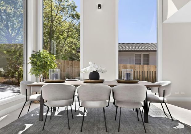 Bright contemporary dining area with a long light-toned dining table surrounded by upholstered dining chairs. Floor-to-ceiling glazing provides views of trees and neighbouring residential buildings, while white walls, a large grey rug, sculptural pendant lighting, and restrained table styling create an airy, refined interior.