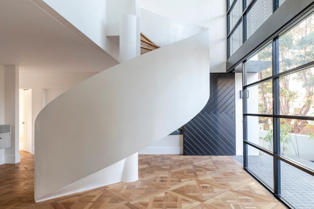 A bright contemporary interior dominated by a sculptural, sweeping white curved staircase or stair balustrade. The space features expansive black-framed floor-to-ceiling glazing, a dark diagonally patterned feature wall, white plastered surfaces, and a richly patterned timber parquet floor. An adjacent hallway and upper-level architectural elements are visible beyond the staircase.