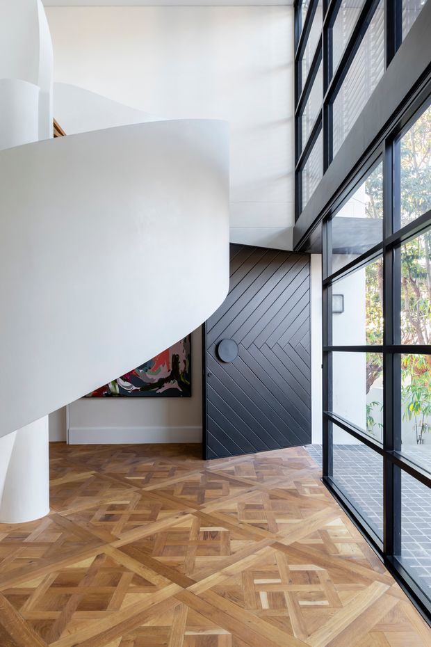A striking contemporary interior entry space with a sculptural white curved staircase or mezzanine form, dramatic double-height volume, full-height black-framed glazing, a dark diagonally panelled door, and intricate patterned timber parquet flooring. Natural light enters through the expansive windows, with a small portion of artwork visible beneath the curved structure.