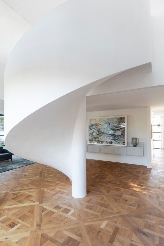 A bright, refined interior featuring a dramatic sculptural curved white staircase or architectural stair enclosure supported by a rounded column. The space has ornate parquet timber flooring, white walls and ceilings, a low built-in grey console with drawers, and an abstract artwork. A glimpse of a dark sofa and adjacent living area is visible at left.