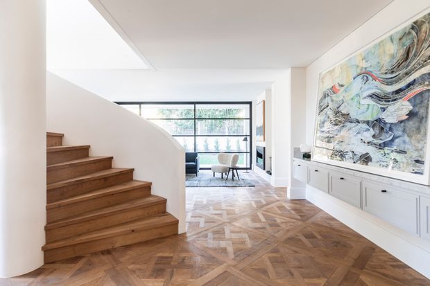 Bright contemporary residential interior showing a timber staircase, patterned parquet flooring, white walls and built-in cabinetry with a large abstract artwork. A glazed living area with seating is visible beyond the stair hall.