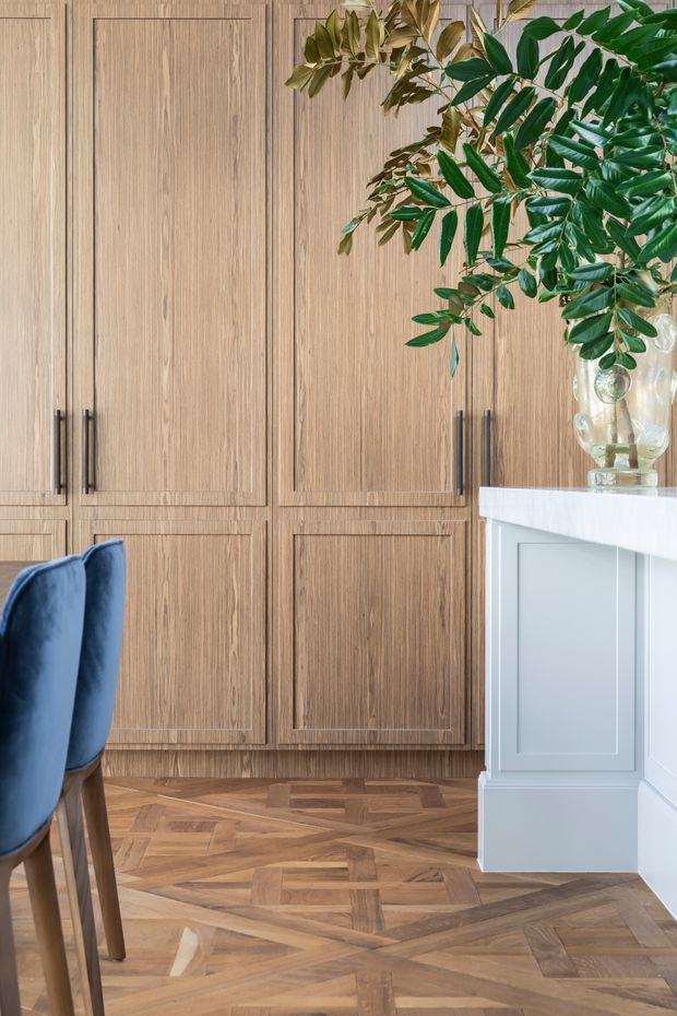 A refined interior detail featuring full-height, warm mid-toned timber cabinetry with framed panel doors and dark vertical handles. The cabinetry sits above a geometric parquet timber floor. At right is a pale blue-grey built-in island or counter with a white stone benchtop, topped by a clear vase of leafy branches. A portion of a navy upholstered dining chair is visible at left. The composition is warm, elegant, and tailored, with a blend of classic joinery and contemporary colour.