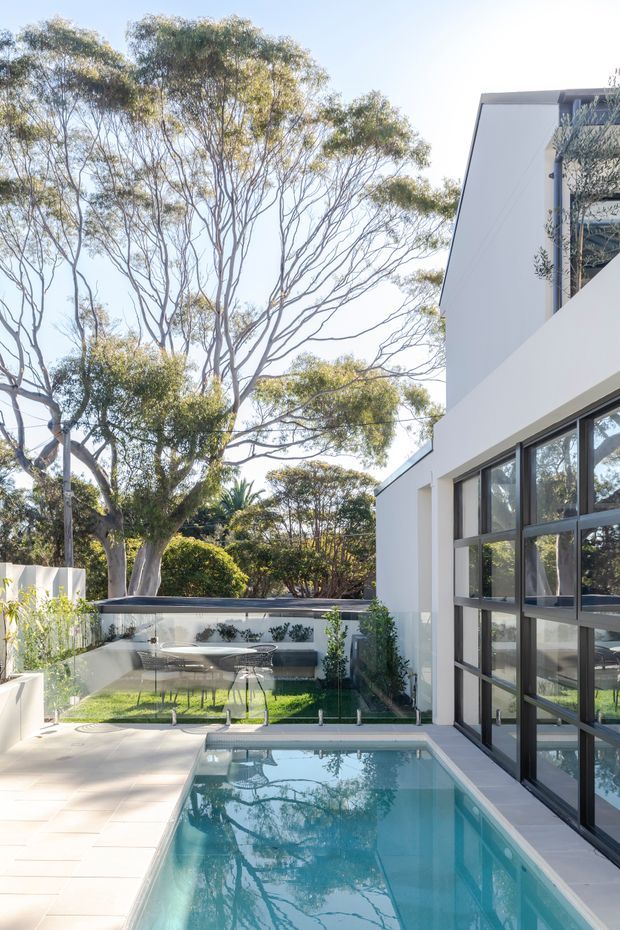 A bright contemporary courtyard viewed from beside a narrow rectangular swimming pool. The white multi-level house has large black-framed glazed doors and windows, with a paved terrace, glass pool fencing, outdoor dining furniture and mature trees surrounding the space.
