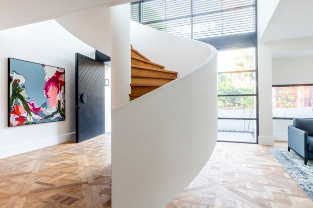 Bright contemporary interior centred on a sculptural curved staircase with warm timber treads and a white plastered balustrade. The space features geometric timber parquet flooring, expansive black-framed glazing with horizontal blinds, a dark pivot entry door, and colourful wall art. A portion of a living area is visible to the right.