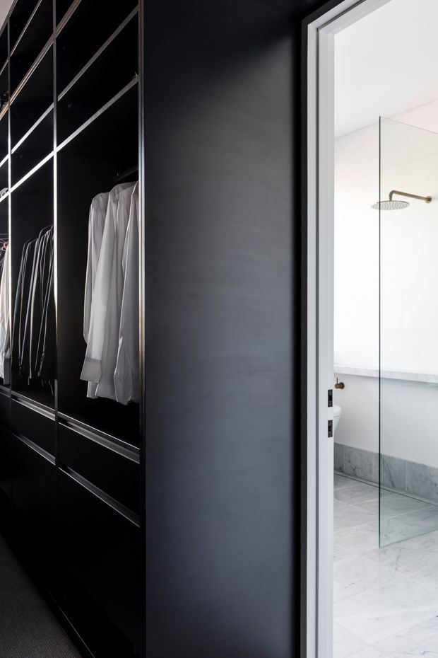 A refined interior view showing a dark built-in wardrobe with open hanging sections and drawers on the left, adjacent to a bright marble bathroom visible through a doorway. The bathroom includes a frameless glass shower screen, large round showerhead, pale stone or marble wall and floor finishes, and warm metallic shower fittings. The composition contrasts the dark wardrobe joinery with the light, high-end bathroom.
