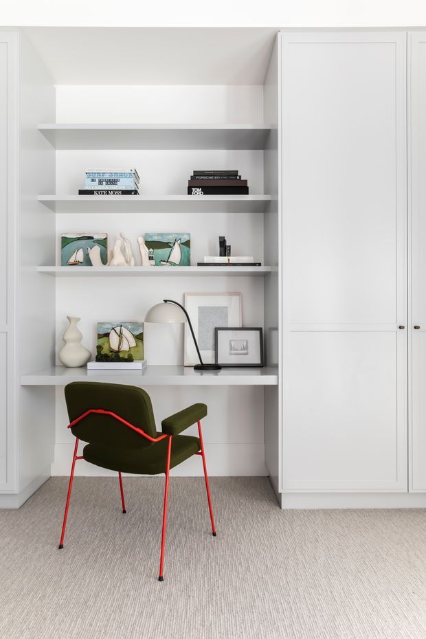 A compact built-in study nook with open white shelving, closed floor-to-ceiling storage cabinetry, and a white desk surface. The shelves display books, framed artwork, small sculptural objects, and a black desk lamp. A dark olive-green lounge chair with a distinctive bright red metal frame sits in front. The space has light neutral tones and textured carpet, creating a clean, curated contemporary interior.