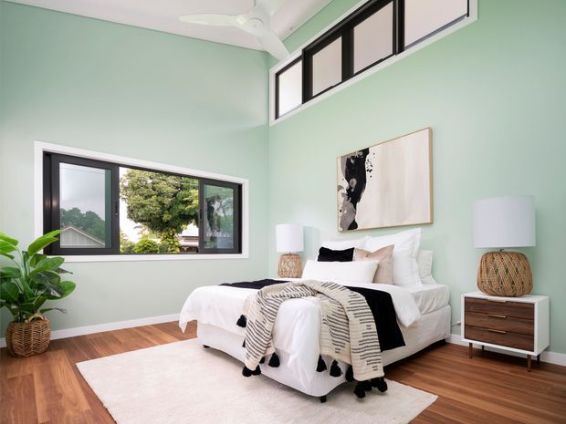 Bright, calm bedroom with pale mint-green walls, warm timber flooring, and high clerestory windows. A white upholstered bed is layered with white, black, and patterned bedding, positioned on a large pale rug. Matching bedside tables with woven lamps flank the bed, while a leafy plant, abstract artwork, ceiling fan, and black-framed windows complete the contemporary interior.