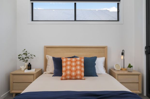 A bright, neatly styled bedroom with a timber bedhead, neutral bedding, matching timber bedside tables, navy cushions and throw, and a vivid orange patterned accent cushion. A high horizontal window provides natural light, with a roof visible outside.
